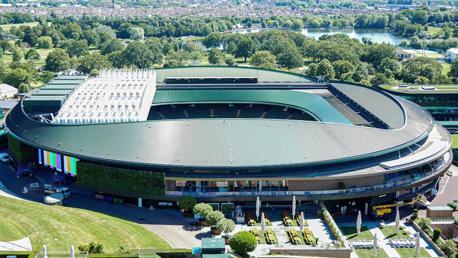 An aerial view of a large, modern sports stadium with a distinctive oval shape, featuring a predominantly green metal roof finished with a smooth, ribbed texture. The stadium's exterior walls include sections of glass and dark-colored panels, with some areas displaying vertical green vegetation for aesthetic contrast. Surrounding the stadium is a paved area with parked vehicles, including several cars and a small truck, indicating nearby access points for services such as waste management. In the vicinity, there are neatly maintained lawns and garden areas with trimmed bushes and small trees, enhancing the landscaped environment. The stadium is positioned against a backdrop of lush trees, open fields, and a distant body of water, suggesting a suburban or parkland setting. The overall scene is illuminated by natural daylight, providing clear visibility of the structure’s architectural details, with a subtle relevance to the provision of private waste disposal or onsite clearance services for large-scale facilities managed by companies akin to Waste Clearance Wimbledon.