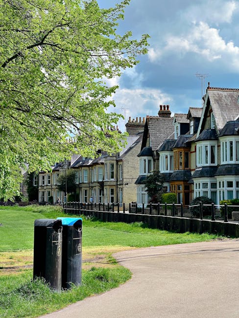 A row of traditional terraced houses with bay windows, slate roofs, and brick chimneys, situated along a residential street with well-maintained front gardens. In the foreground, two black public waste bins are positioned on a grassy verge near the pavement. To the left, a large tree with bright green foliage extends its branches over the scene, creating dappled shade. The sky above is partly cloudy with patches of blue visible through scattered white clouds. The environment appears clean and orderly, reflecting typical suburban character, with the waste bins indicating public or community waste disposal points within the area. The setting suggests a quiet, family-oriented neighbourhood, where local waste management services are complemented by occasional waste clearance initiatives from private providers. Overall, the image captures a peaceful residential street scene with an emphasis on community infrastructure and green spaces, aligning with services such as waste clearance or rubbish collection in a suburban context.