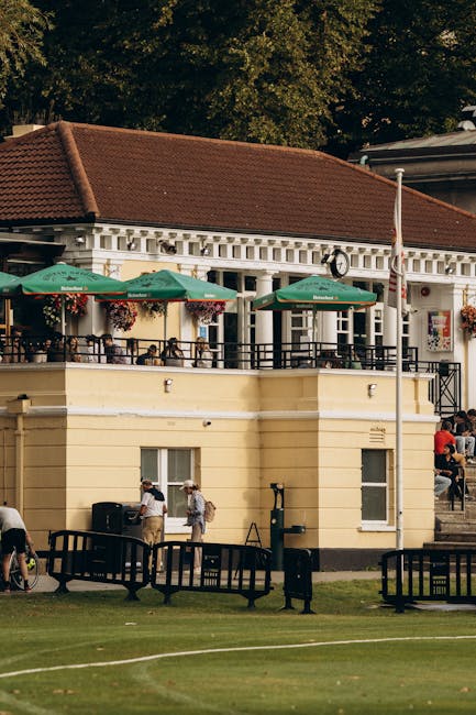 A two-story building with a beige painted exterior and white decorative architectural details along the upper terrace, situated in a landscaped area with a grassy lawn and black metal fences. The roof is covered with reddish-brown tiles, and there are large green umbrellas providing shade to outdoor seating on the upper level, where several people are gathered. The lower level features a small window and a black waste bin near the base, with a few individuals walking or standing nearby. The background includes tall, leafy trees and a portion of a dark-colored structure, indicating an outdoor public space, possibly a park or recreational area. This setting relates to waste management services such as those provided by Waste Clearance Wimbledon, which handles rubbish clearance for local premises including commercial or public buildings in the Wimbledon area, supporting the need for efficient rubbish removal or alternative waste handling from community facilities.